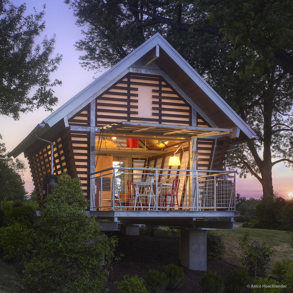 A modern house with a sloped roof showcases a welcoming interior, illuminated by warm lights, surrounded by trees and shrubs at sunset.