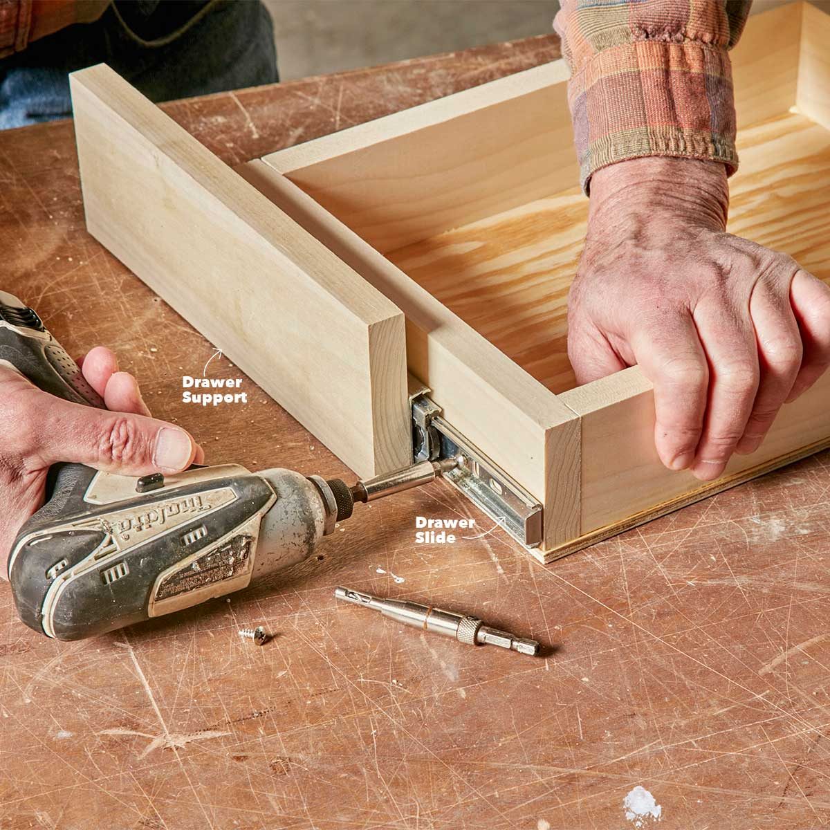 A hand uses a drill to attach a drawer slide to a wooden drawer. The workspace features a wooden table and various tools nearby.