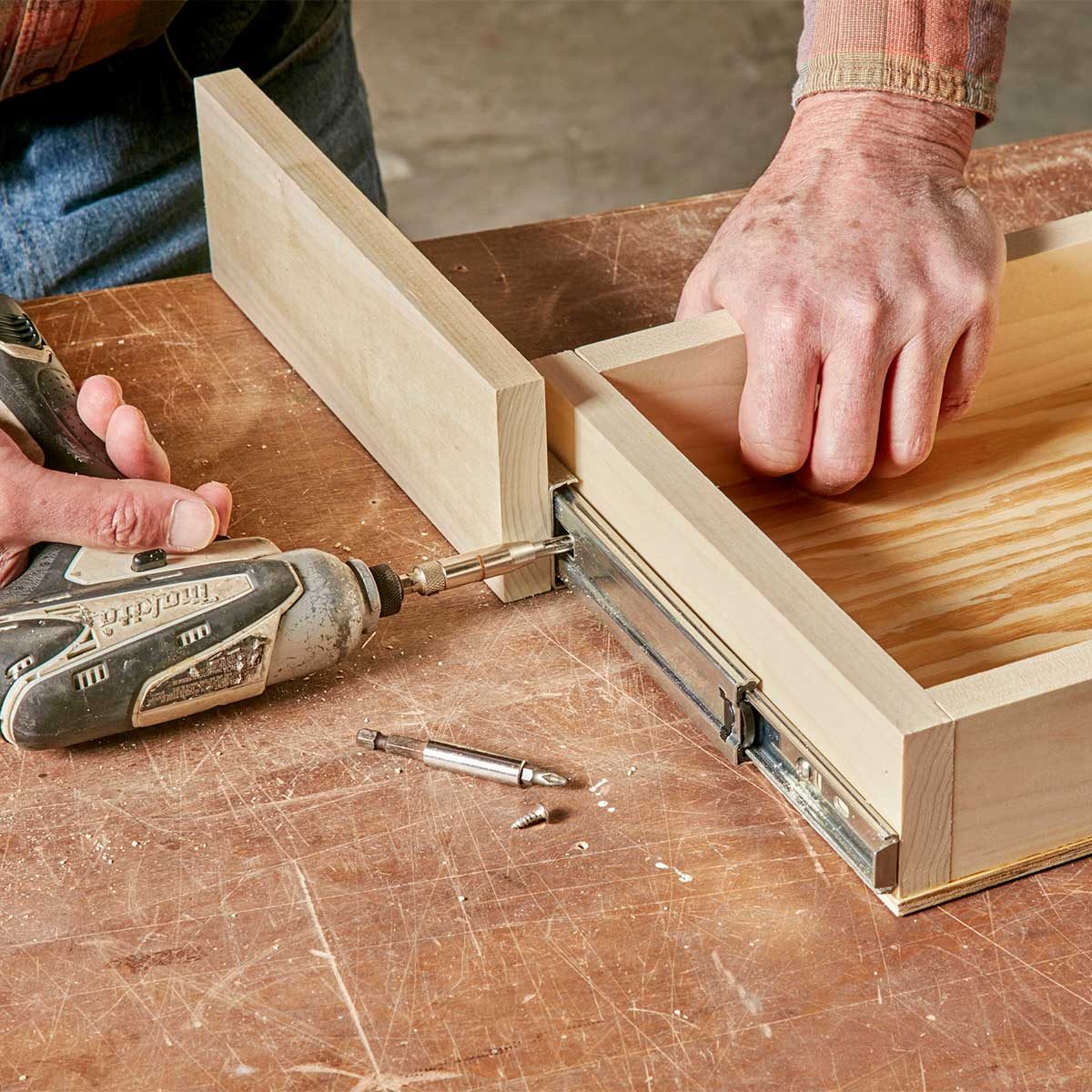 A person uses a power drill to install a sliding mechanism on a wooden drawer, surrounded by tools and a wooden work surface.