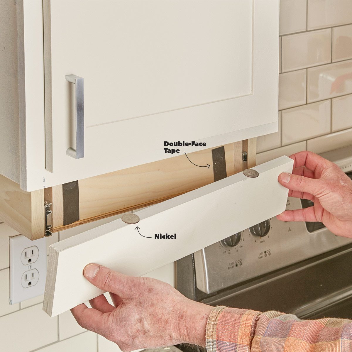 A hand holds a cabinet's false front, adhering it with double-face tape above a nickel, positioned near kitchen appliances and tiled walls.