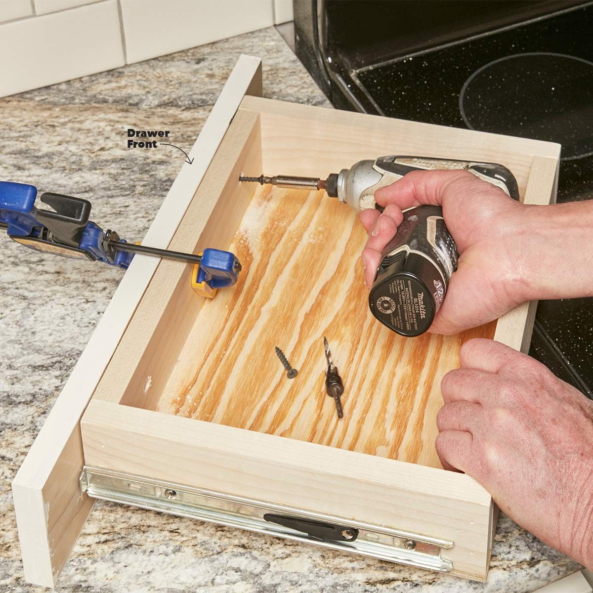 A hand holds a drill to attach a drawer front, while a clamp secures the drawer on a granite countertop with scattered screws nearby.
