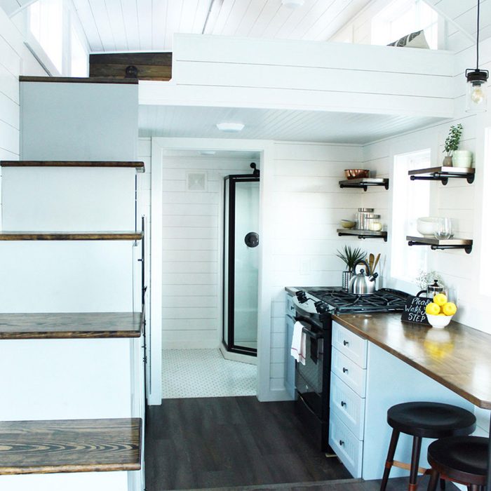 A small, modern kitchen features white walls, a stove, and shelves. Stairs lead to a loft, while a shower is visible in the adjacent space.