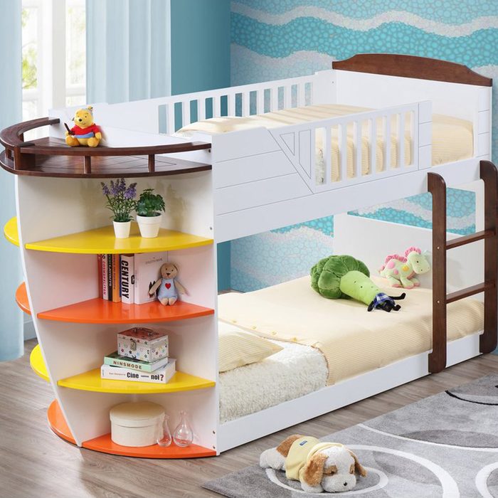 A multilevel bed with a storage bookshelf on the side, featuring soft toys, against a blue wall pattern in a room with wooden flooring.