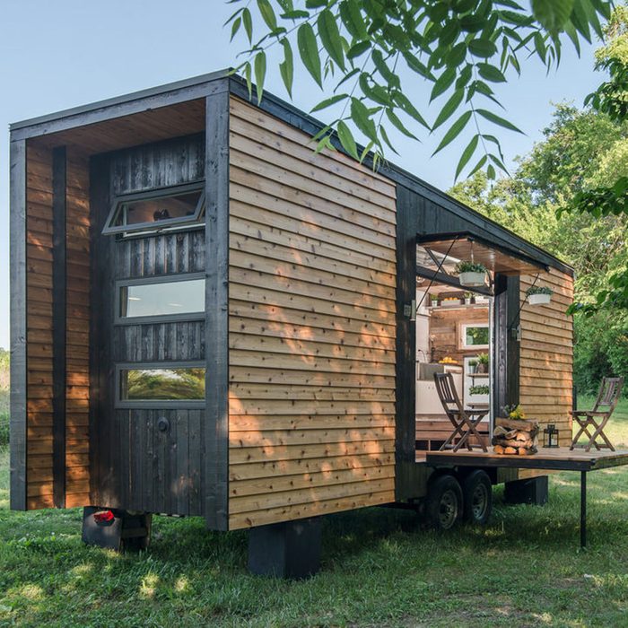 A tiny wooden house on wheels, with large windows, features an open interior and a small deck, surrounded by greenery under a clear blue sky.