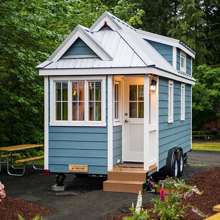 A small, blue tiny house with a metal roof sits on wheels. It features multiple windows and a white door, surrounded by greenery and picnic seating.