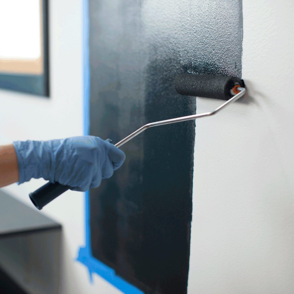 A paint roller, held by a gloved hand, applies dark paint to a wall with blue painter's tape nearby, in a bright interior setting.