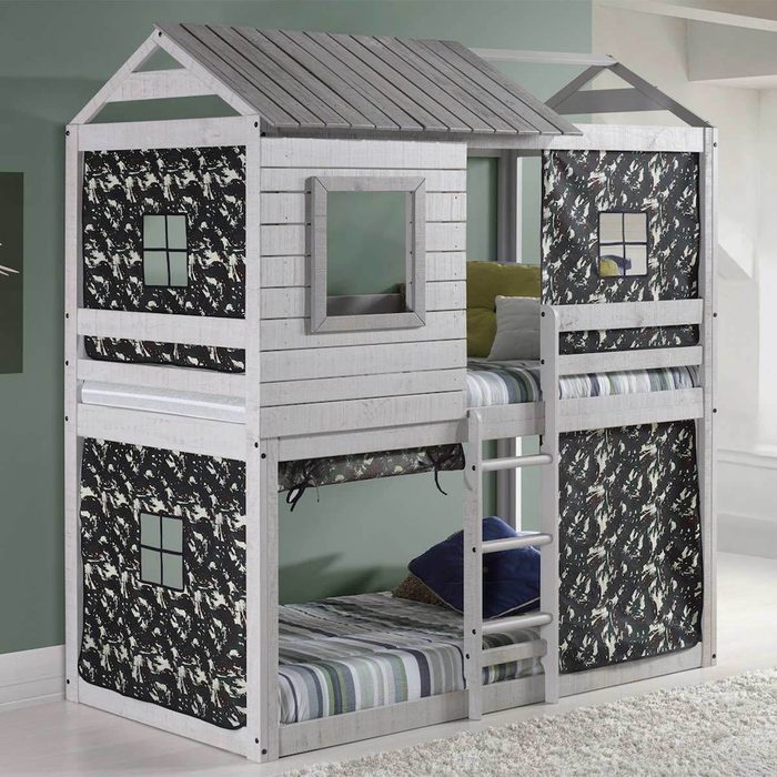 A wooden bunk bed resembling a house features a ladder, green walls, and striped bedding, with camouflage-patterned fabric for privacy on the lower level.