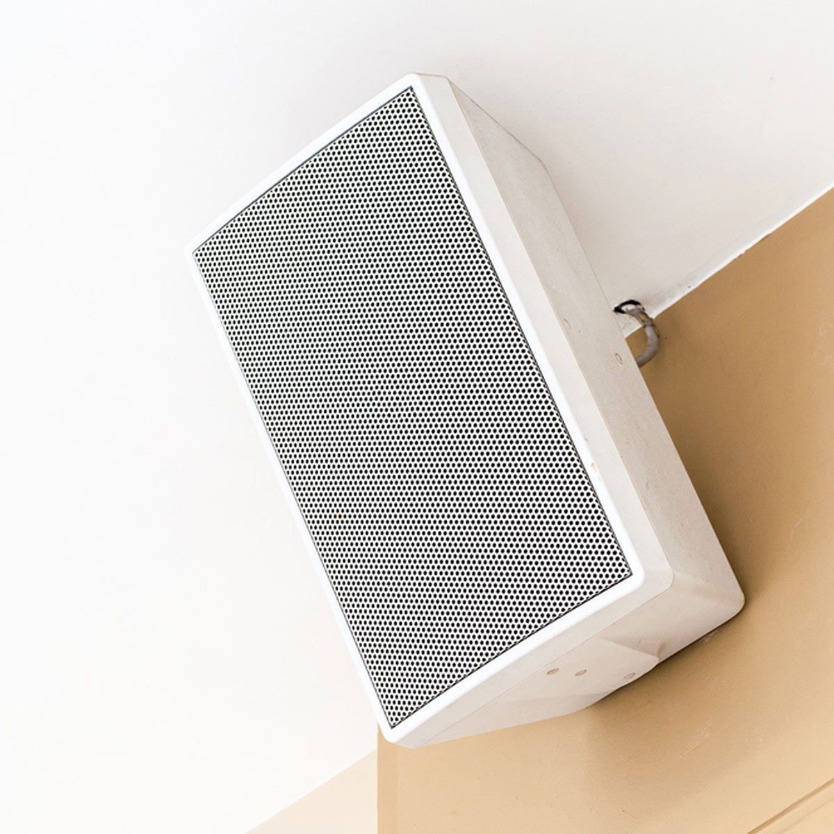 A white ceiling-mounted speaker emits sound, situated at the corner where a white wall meets a beige wall, blending into the indoor environment.