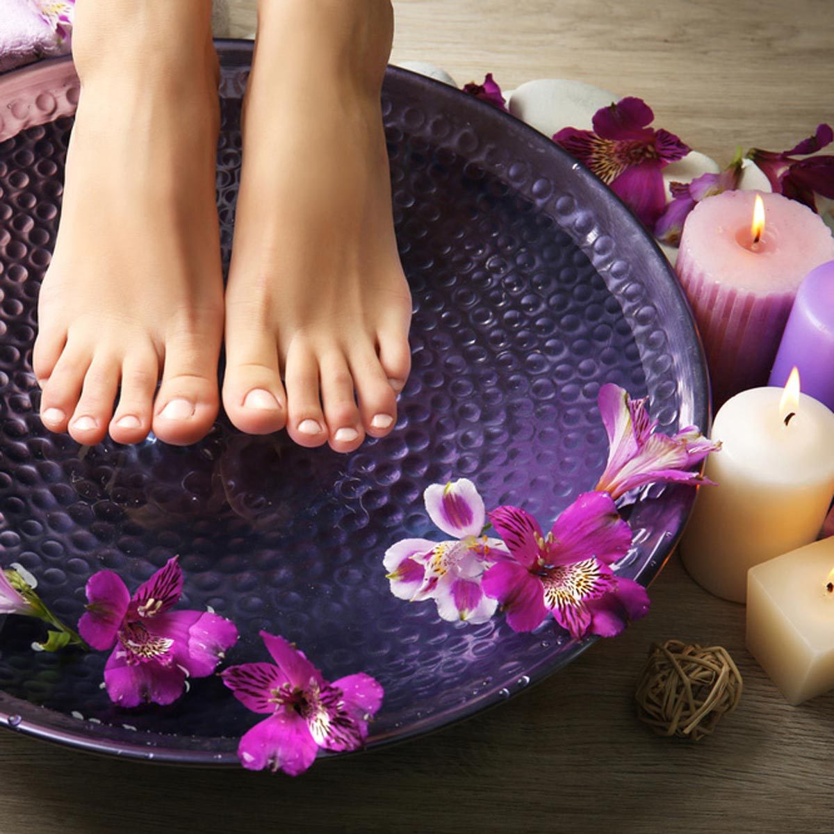 Feet rest in a purple bowl of water, surrounded by flowers and lit candles, creating a relaxing spa-like atmosphere.