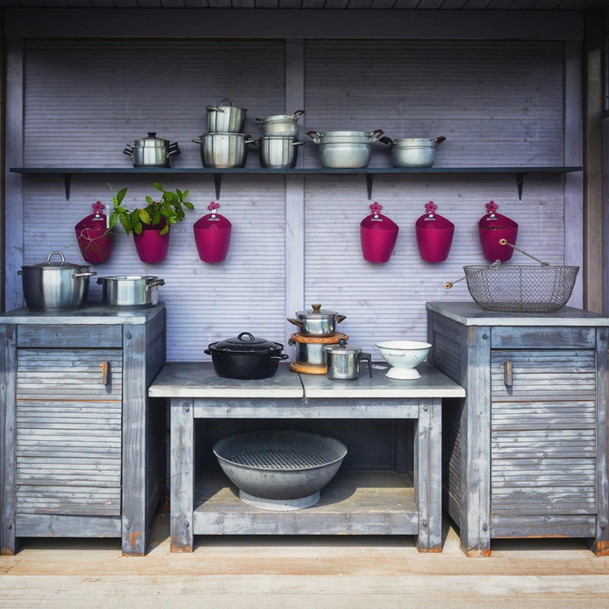 Metal pots and pans are arranged on shelves and surfaces within a rustic kitchen space, accented by colorful hanging planters.