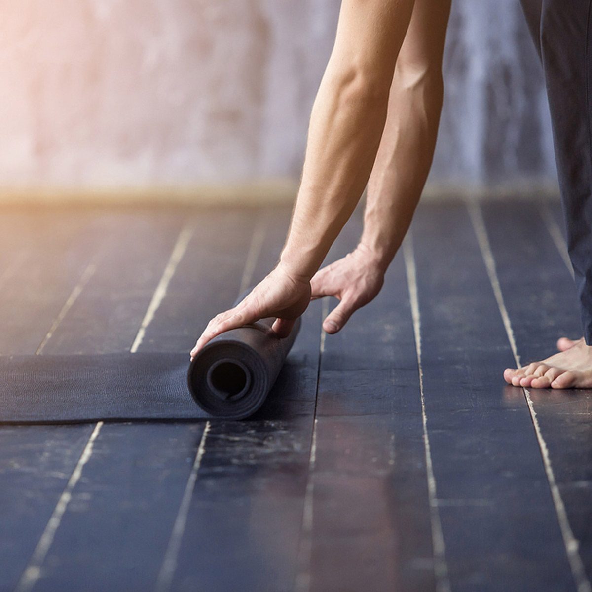 A hand rolls out a black yoga mat on a wooden floor, preparing for exercise in a well-lit, minimalist space with a textured wall.