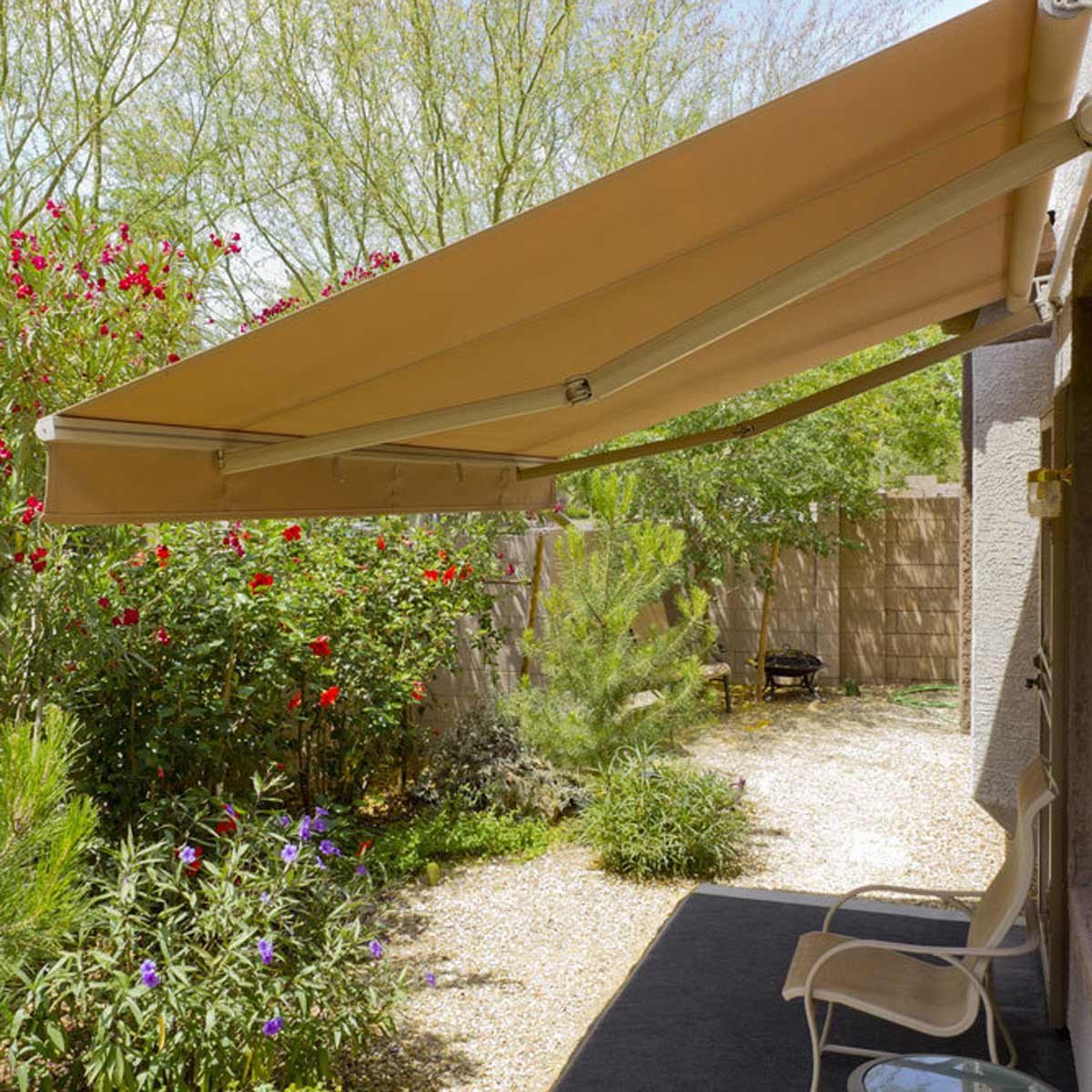 An awning extends over a stone pathway, providing shade near flowering plants in a garden, with a chair positioned for outdoor relaxation.