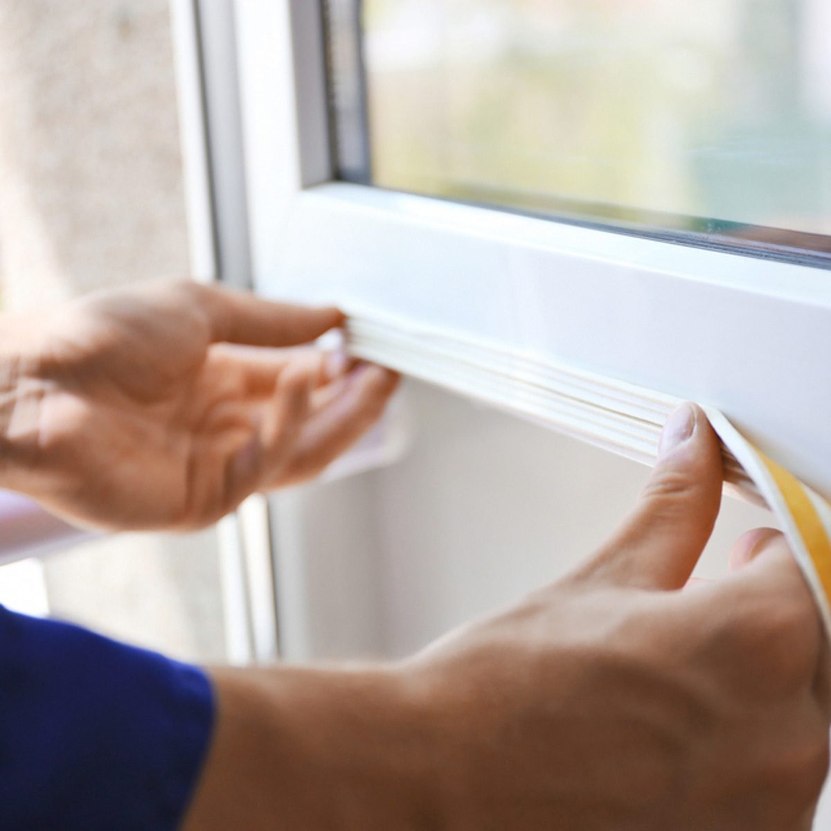 A person is applying adhesive tape to the edge of a window frame, focusing on creating a seal in a bright, indoor environment.