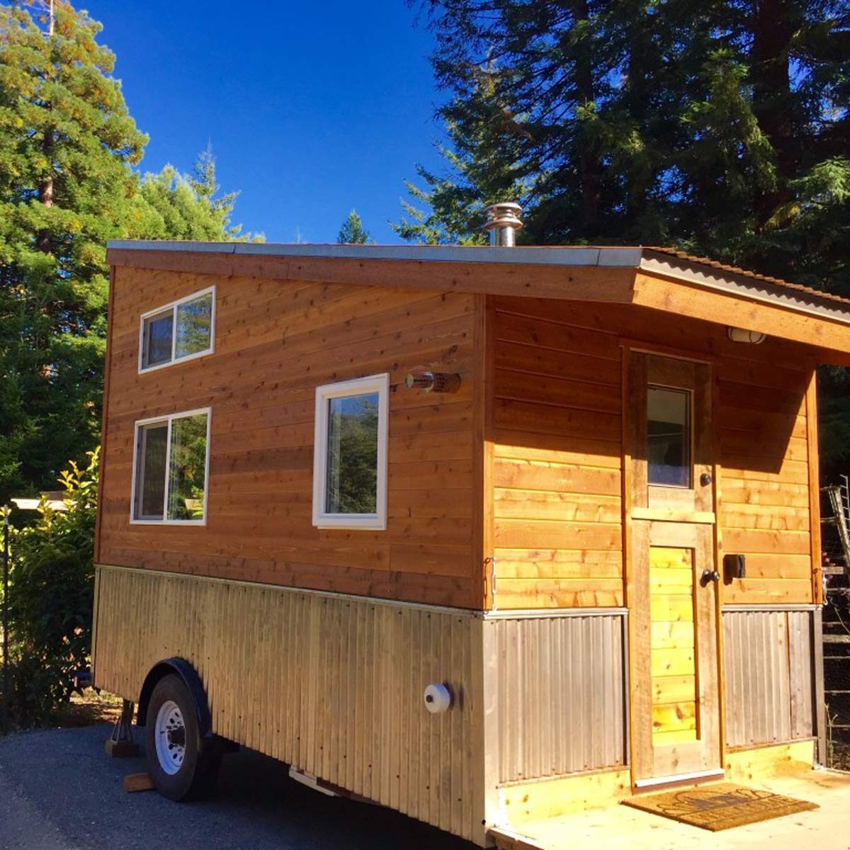 A wooden tiny house on wheels sits under a clear blue sky, surrounded by lush green trees, featuring multiple windows and a front door.