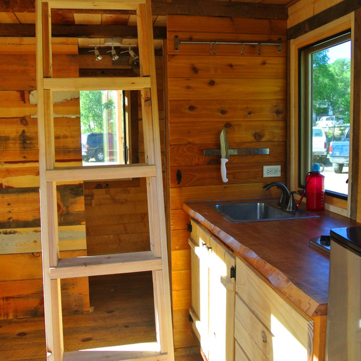 A wooden ladder leans against a wall, while a kitchen area features a sink, countertop, and a knife on a magnetic strip in a small, rustic space.