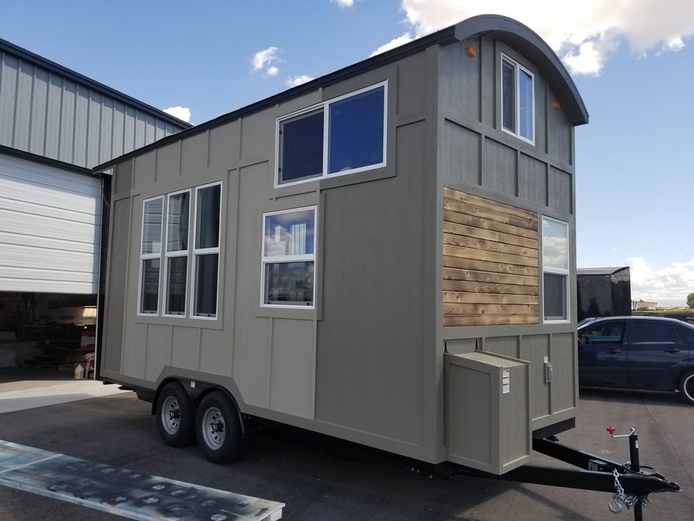 A small trailer home features multiple windows and a wooden accent wall, parked beside a large garage in a clear sky setting.