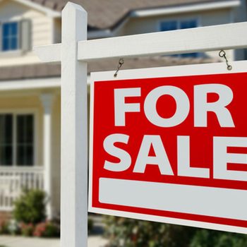 A red "FOR SALE" sign is displayed outside a house, indicating it is available for purchase, surrounded by a landscaped yard and a sunny atmosphere.