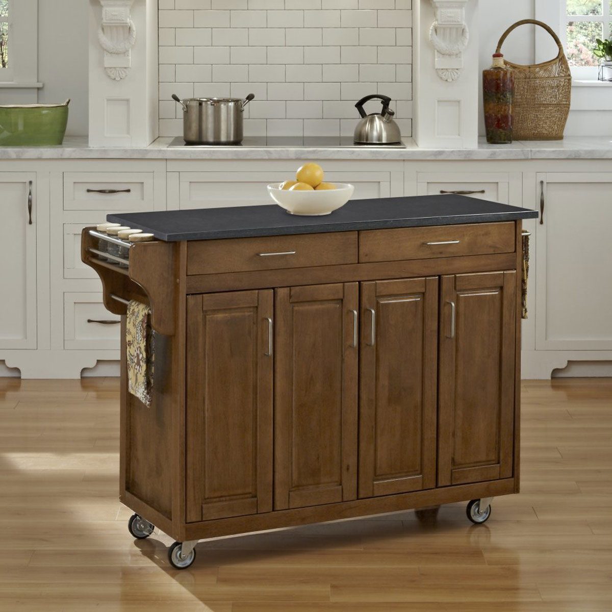 A wooden kitchen island on wheels supports a bowl of lemons, situated in a bright kitchen with white cabinets and a marble countertop in the background.