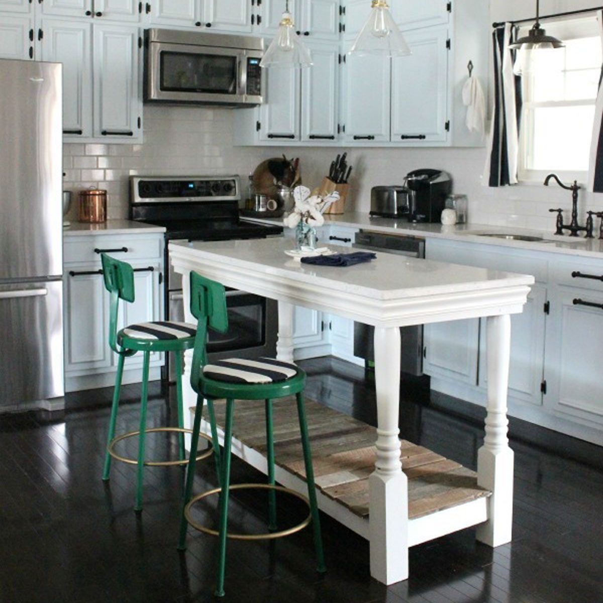 A white kitchen island with two green bar stools stands in a modern kitchen, surrounded by light blue cabinets and stainless steel appliances.