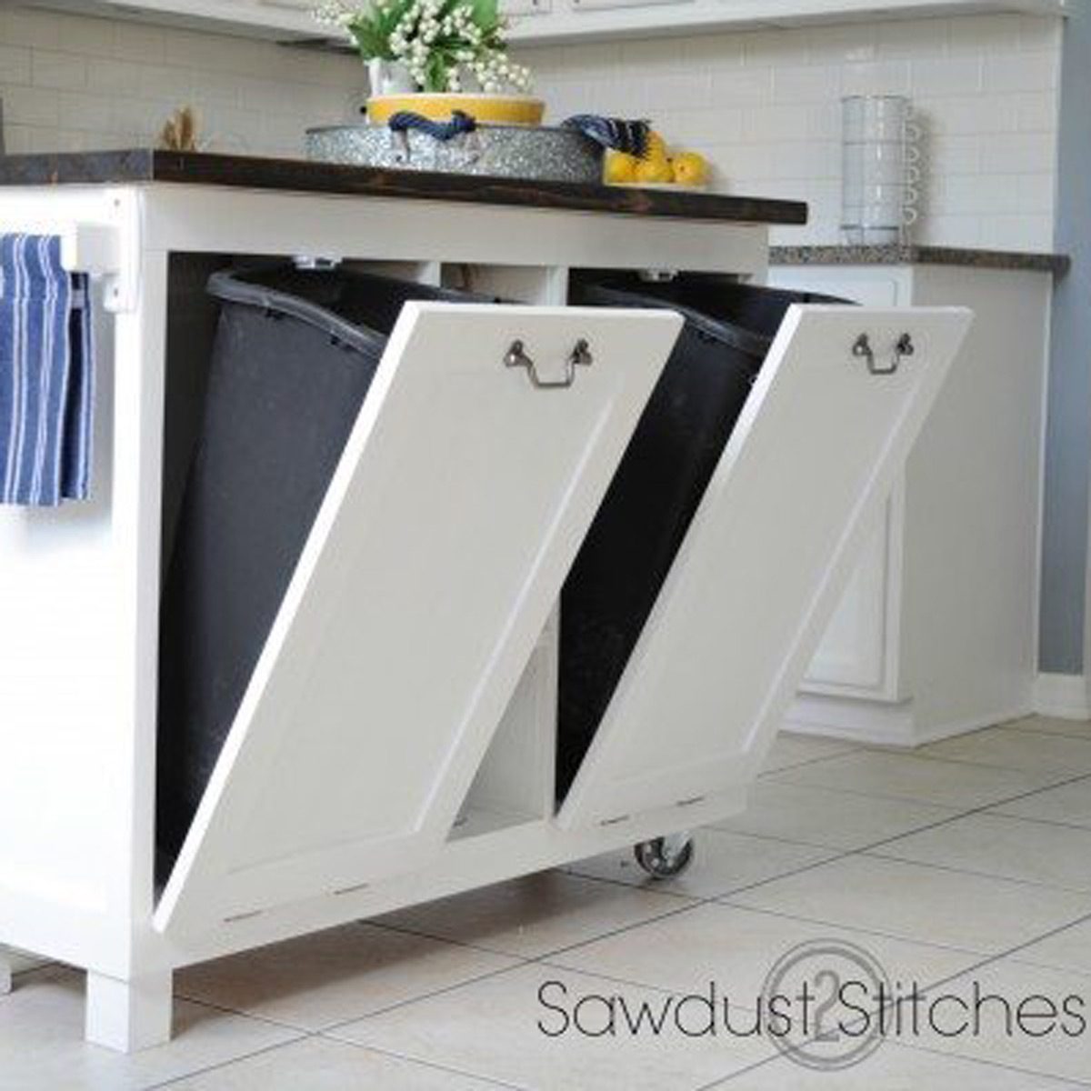 A double-door cabinet reveals two black trash bins, positioned beneath a wooden countertop in a bright kitchen. A towel hangs nearby.
