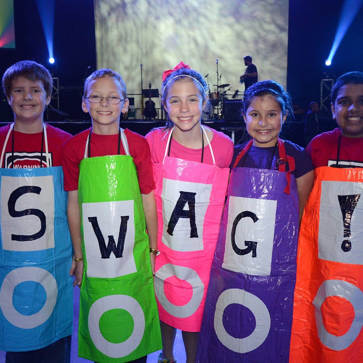 Five children, wearing colorful aprons with large letters, stand smiling on a stage lit with vibrant lights, showcasing the word "SWAG."