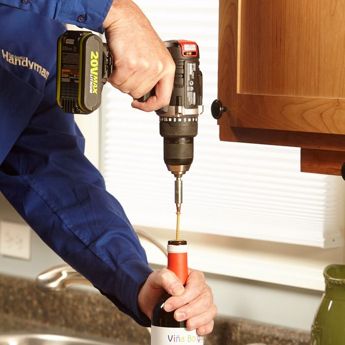 A person uses a drill to extract a cork from a wine bottle while standing at a countertop in a well-lit kitchen.