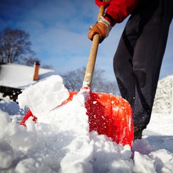A person uses a red shovel to lift snow, creating a plume of snowflakes in a bright outdoor setting with a clear sky and trees in the background.