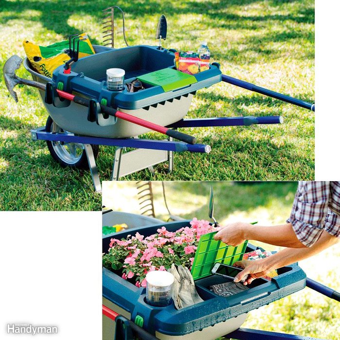 A wheelbarrow filled with gardening tools and supplies rests on grass while a person removes a green tray from it, revealing pink flowers.