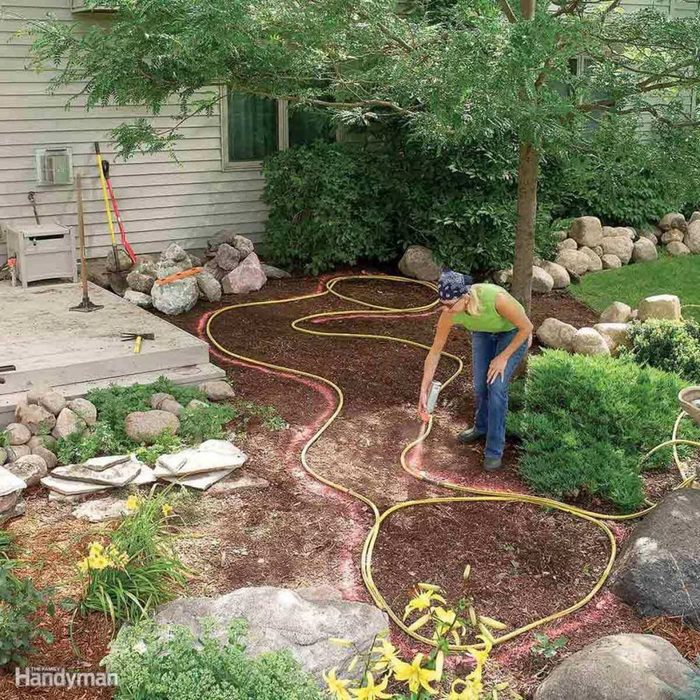 A person waters garden soil using a hose, while surrounded by plants, rocks, and a house structure in a landscaped area.