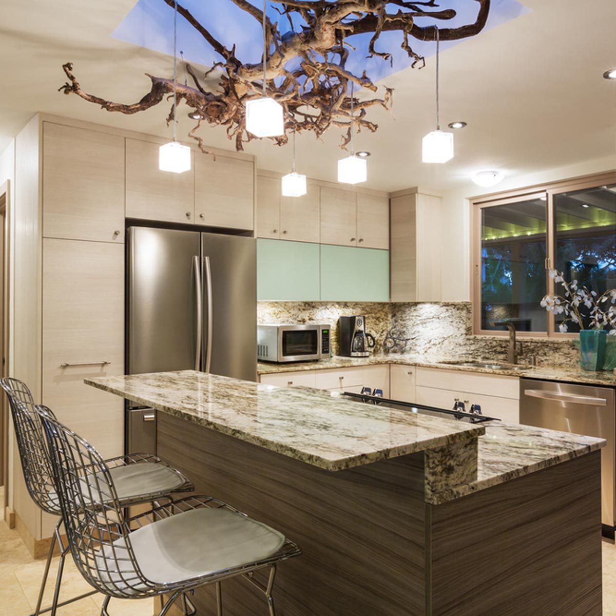 A modern kitchen features a granite island with bar stools. Pendant lights hang from a woody ceiling, illuminating stainless steel appliances and cabinetry.