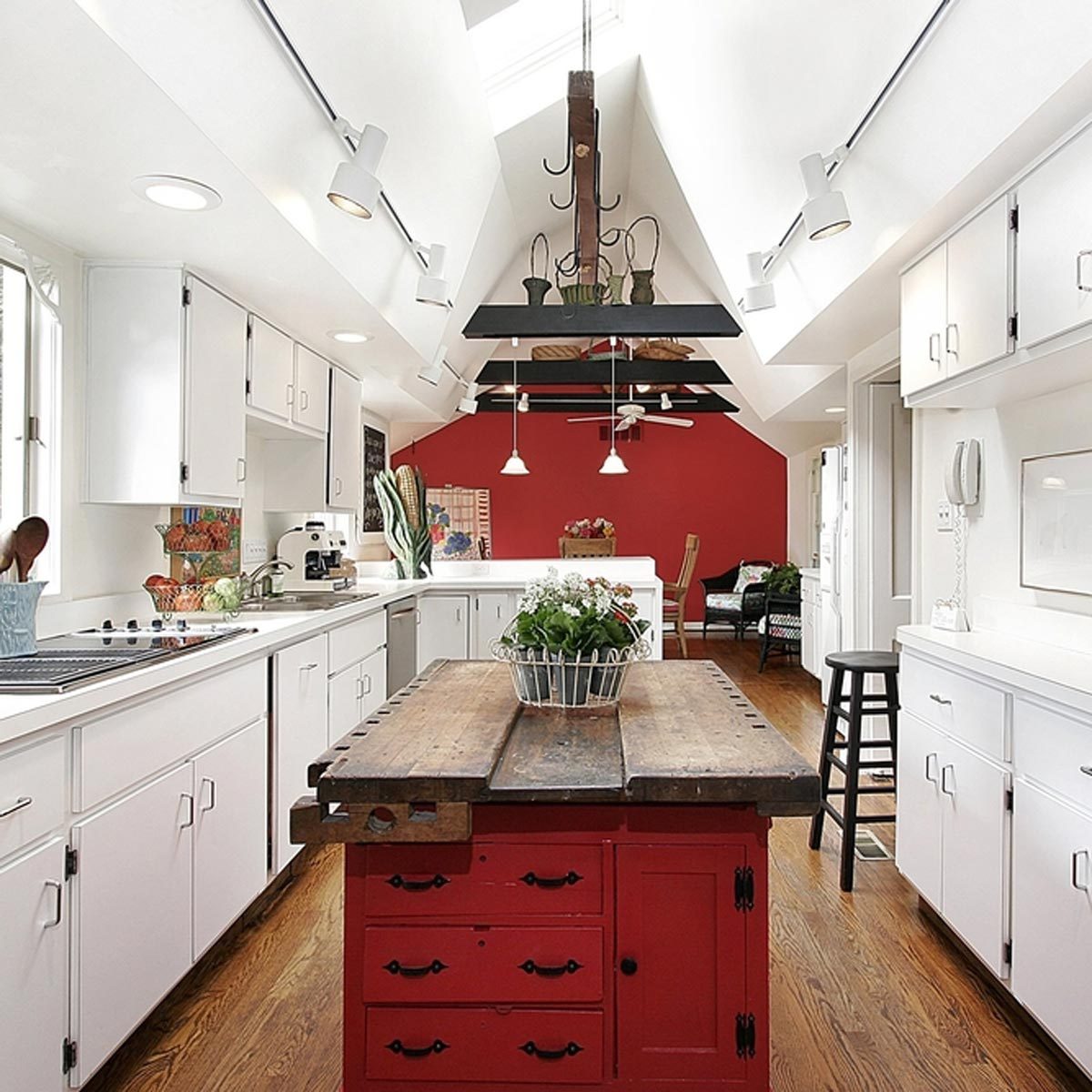 A red kitchen island with plants contrasts against white cabinets and a sloped ceiling, illuminated by modern lights in a bright, spacious kitchen.