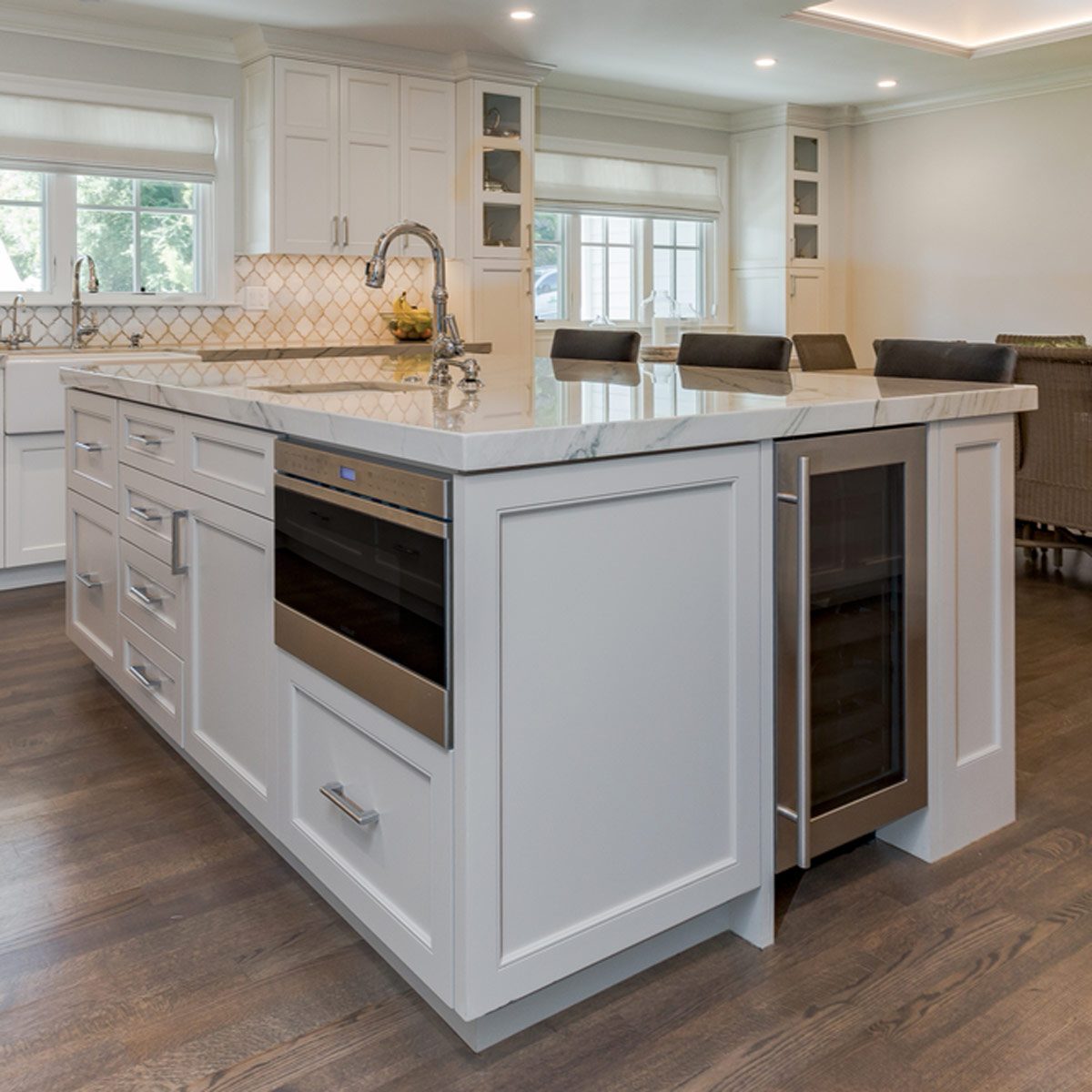 A white kitchen island with a marble countertop displays a sink and wine cooler, surrounded by light-colored cabinetry and wooden floors in a modern kitchen.