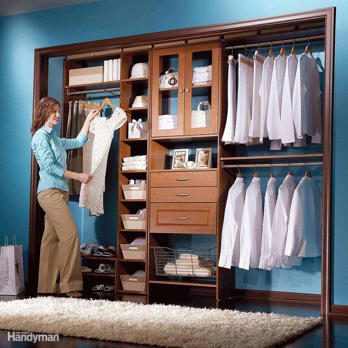 A woman holds a dress in a well-organized closet, featuring shelves, drawers, and hanging clothes, against a blue wall with a plush rug below.