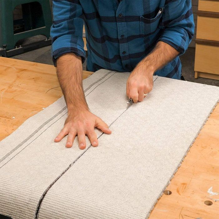 A person is cutting fabric on a wooden workbench, using a utility knife with both hands, surrounded by a workshop environment.