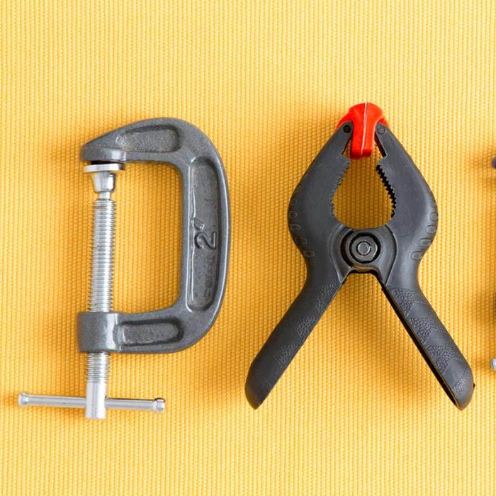 Two hand tools, a C-clamp and a spring clamp, rest on a textured yellow surface, suggesting usage for securing objects together.