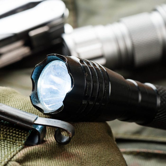 A black flashlight emits light, resting on a green fabric surface alongside a multi-tool device in a rugged, outdoor context.