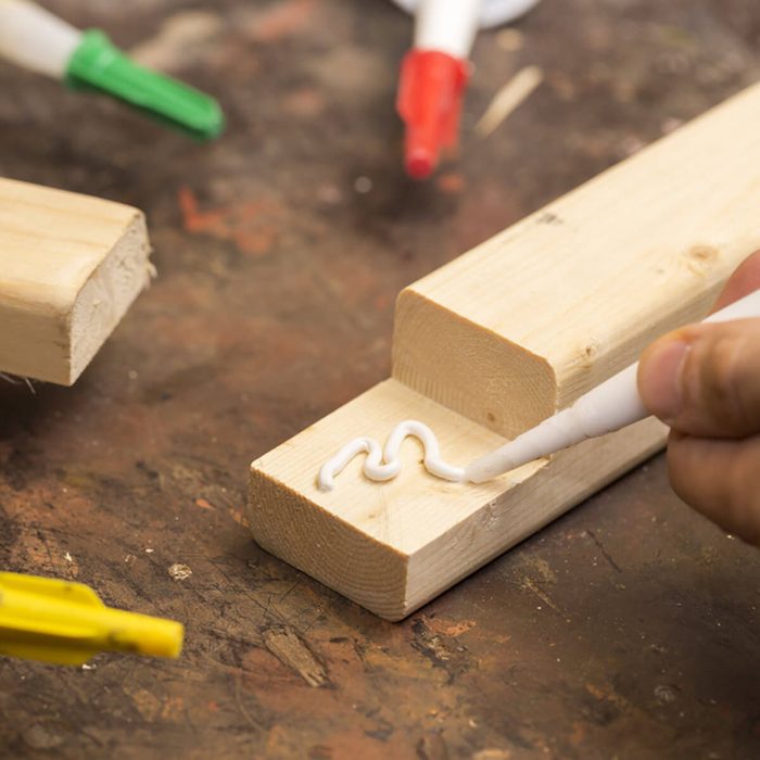 A hand applies white adhesive in a zigzag pattern onto a wooden plank, surrounded by crafting tools on a textured work surface.