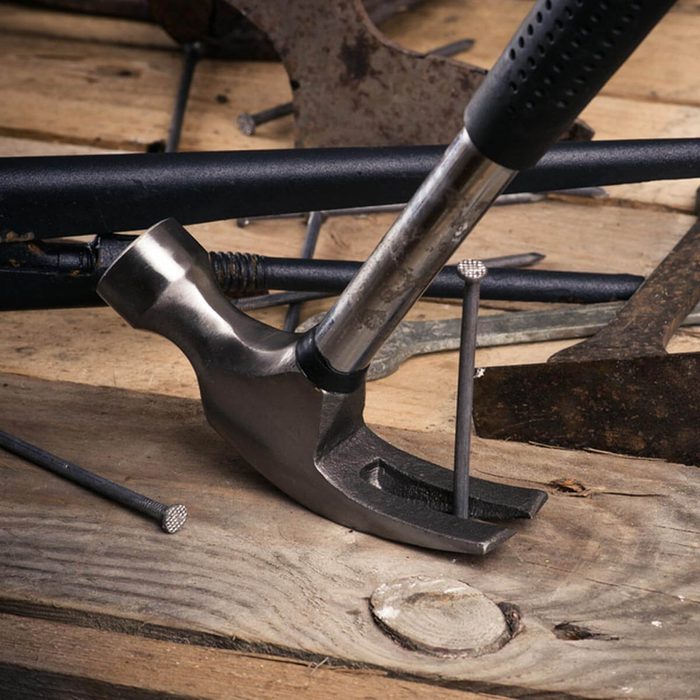 A hammer strikes a nail embedded in wooden planks, surrounded by various tools and scattered metal objects, indicating a workspace or workshop setting.