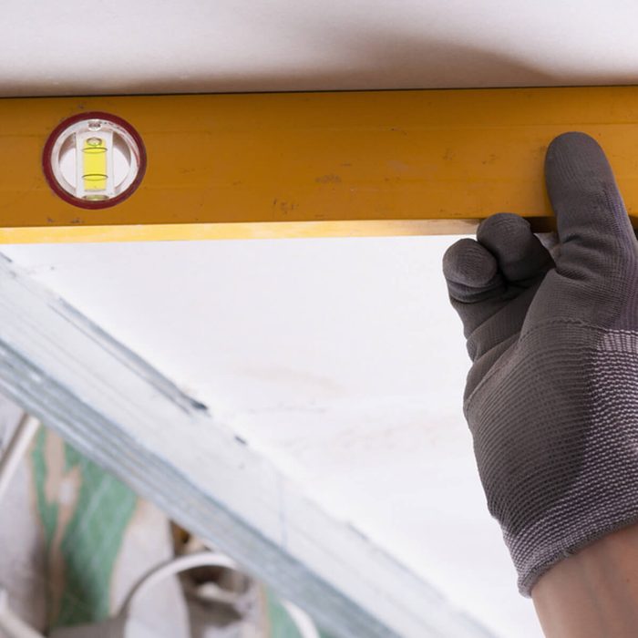 A gloved hand holds a yellow level against a ceiling, ensuring it is straight in a construction environment with visible materials and tools.