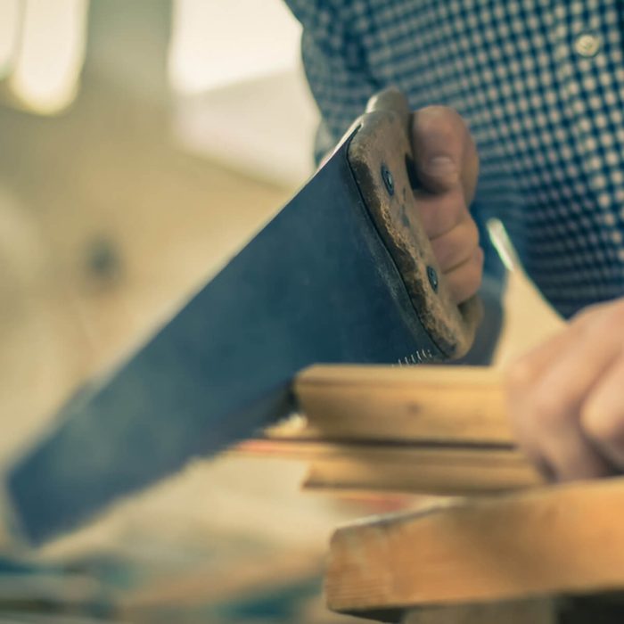 A hand grips a saw, cutting through a piece of wood in a workshop with blurred background details, indicating a focused woodworking activity.