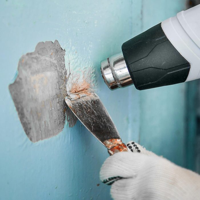 A hand with a gloved grip holds a metal scraper, removing paint from a wall while a heat gun is aimed at the surface to assist.