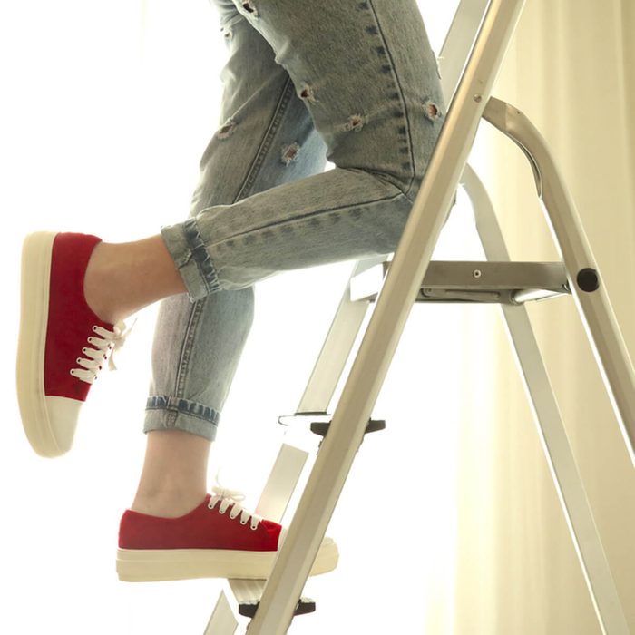 A foot in a red sneaker steps up a ladder, with denim jeans showing visible rips, against a softly lit, bright background.