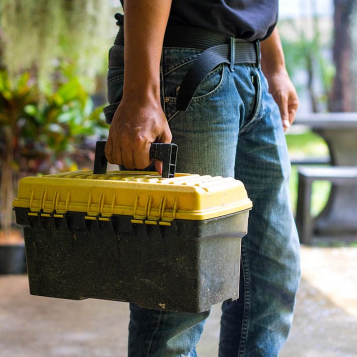A person holds a yellow and black toolbox, standing outdoors amidst greenery and stone benches, suggesting preparation for a task or project.