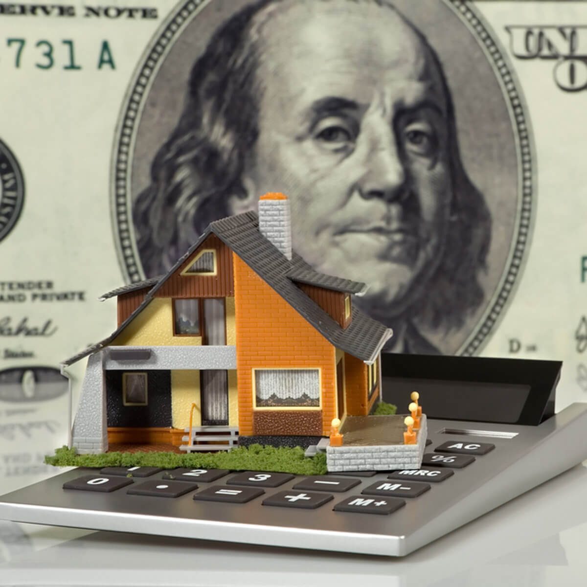 A miniature house sits atop a calculator, symbolizing financial calculation, with a backdrop of a hundred-dollar bill, indicating the context of real estate and money.