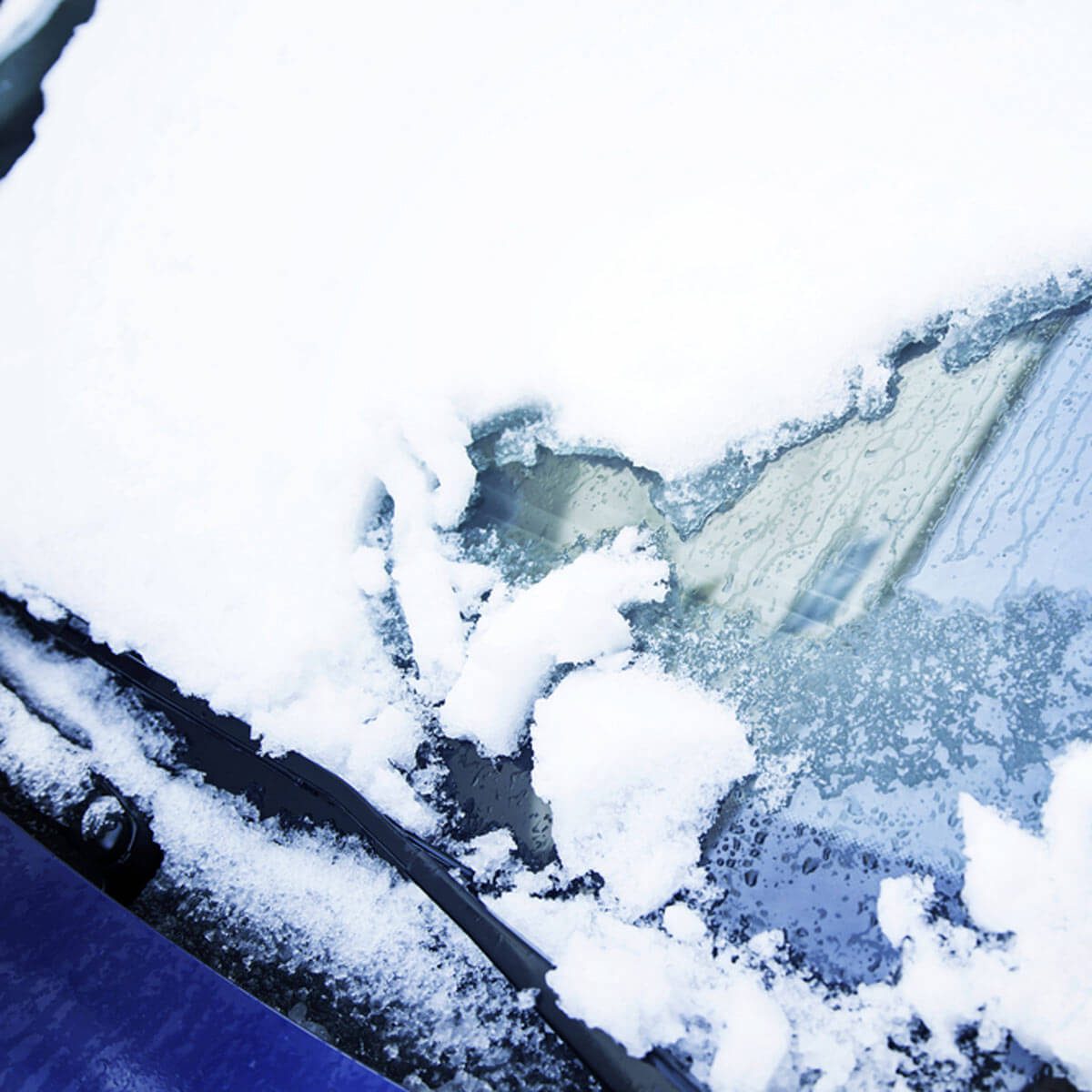 A car's windshield is partially cleared of fresh snow, revealing patches of glass beneath while the surrounding area remains covered in white snow.