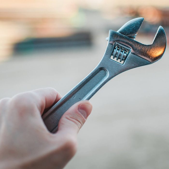 A hand holds a shiny adjustable wrench, poised for use in a construction or repair setting with a blurred background of equipment and surfaces.