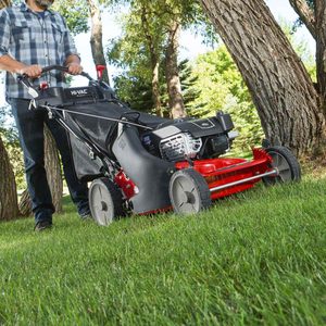 A person operates a red lawn mower on lush green grass, surrounded by trees in a sunny outdoor setting.