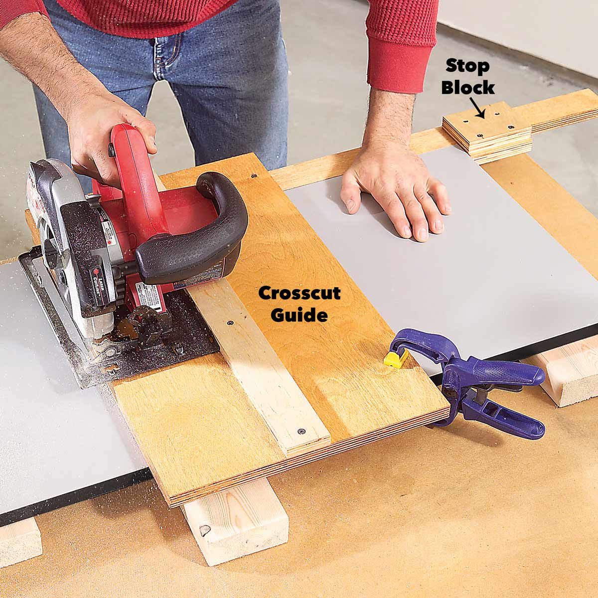 A person uses a circular saw to cut a sheet of material on a wooden surface, guided by a crosscut guide and secured with a clamp.