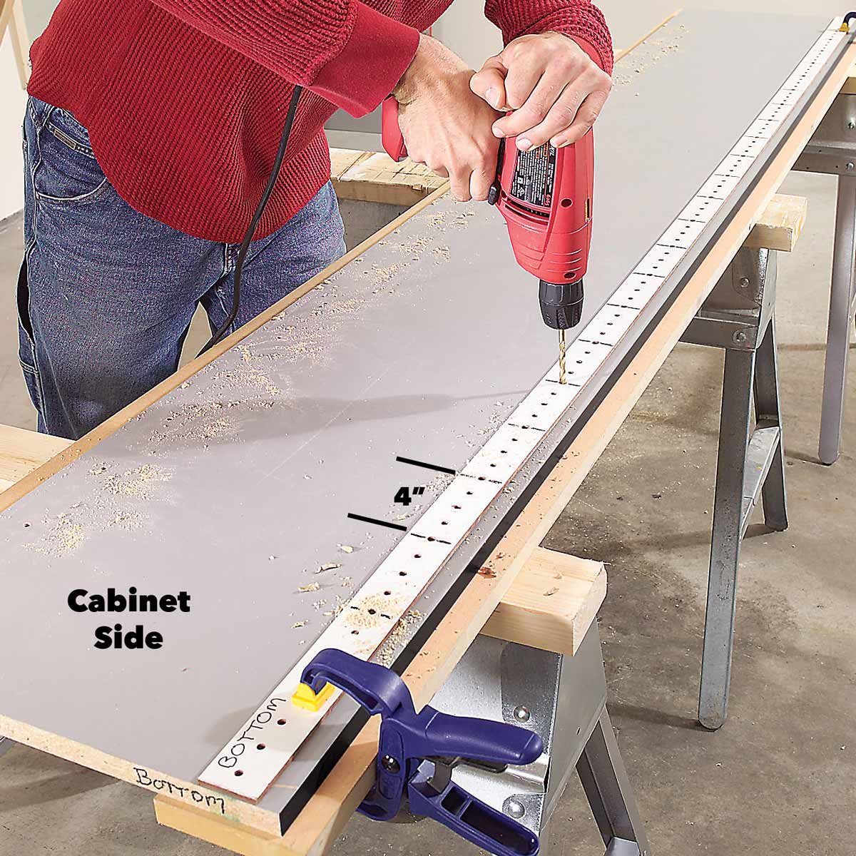 A person drills holes into a cabinet side, secured on a workbench, surrounded by sawdust in a workshop environment.