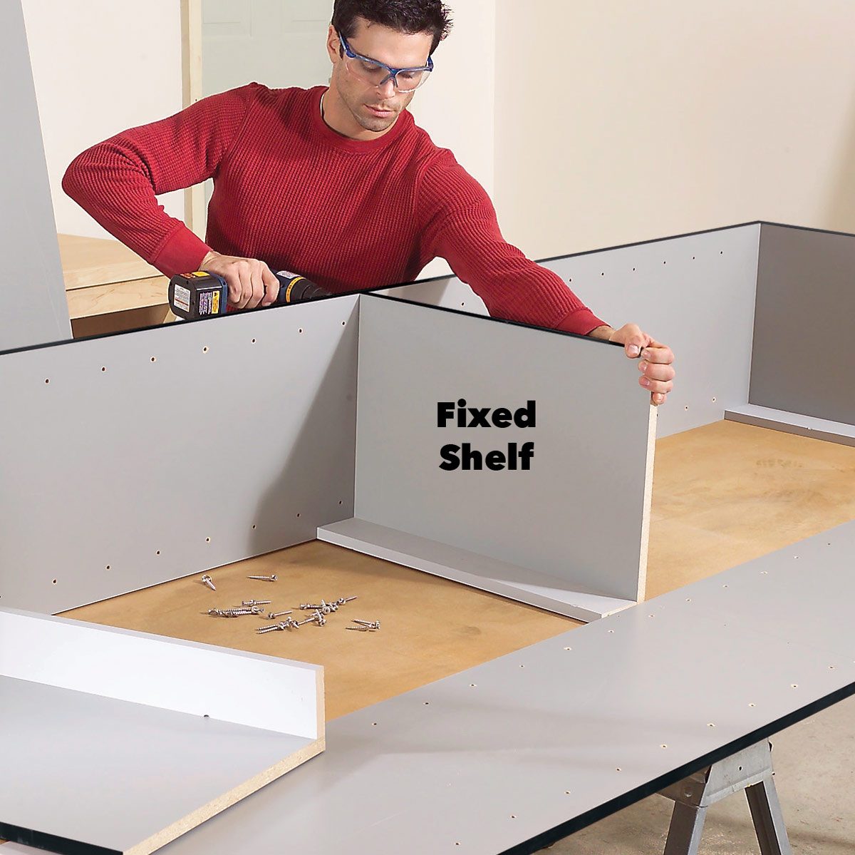 A man in a red sweater uses a power drill to install a fixed shelf in an unfinished wooden structure surrounded by screws and tools.